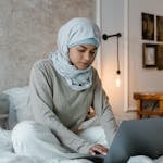 A young Muslim woman wearing a hijab works on her laptop in a stylish, contemporary bedroom setting.